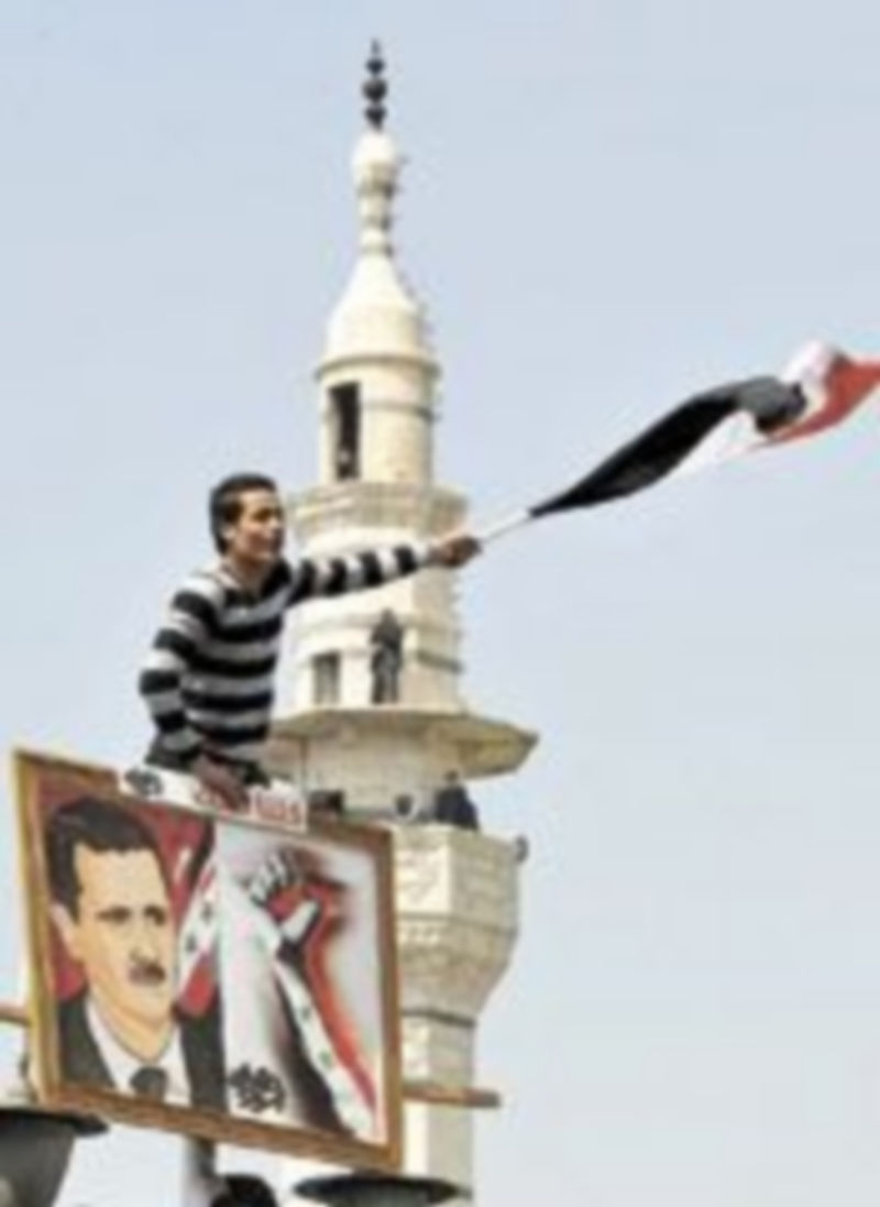 Un hombre ondea la bandera nacional al lado de una imagen de Al Assad. /Reuters