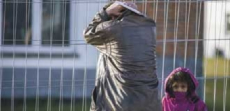 Un hombre y una niña esperan junto a la valla de un centro de registros de refugiados montado en un cuartel militar en una ciudad militar. / EFE