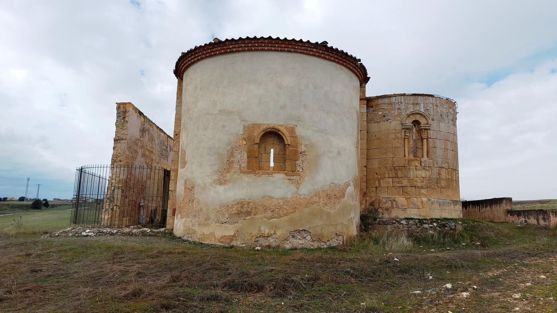 Exterior de la cabecera de la ermita desde el este. Se conservan los ábsides de la capilla mayor y nave del Evangelio (en sillería). La reja de la izquierda muestra el perímetro semicircular del desaparecido ábside del lado de la Epístola. Exterior de la cabecera de la ermita desde el este. Se conservan los ábsides de la capilla mayor y nave del Evangelio (en sillería). La reja de la izquierda muestra el perímetro semicircular del desaparecido ábside del lado de la Epístola.