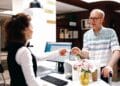 Detailed image shows caucasian retired senior man receiving access card from female receptionist at registration counter. Elderly male tourist giving room key to employee at front desk in hotel lobby.