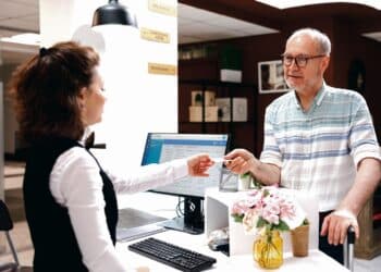Detailed image shows caucasian retired senior man receiving access card from female receptionist at registration counter. Elderly male tourist giving room key to employee at front desk in hotel lobby.
