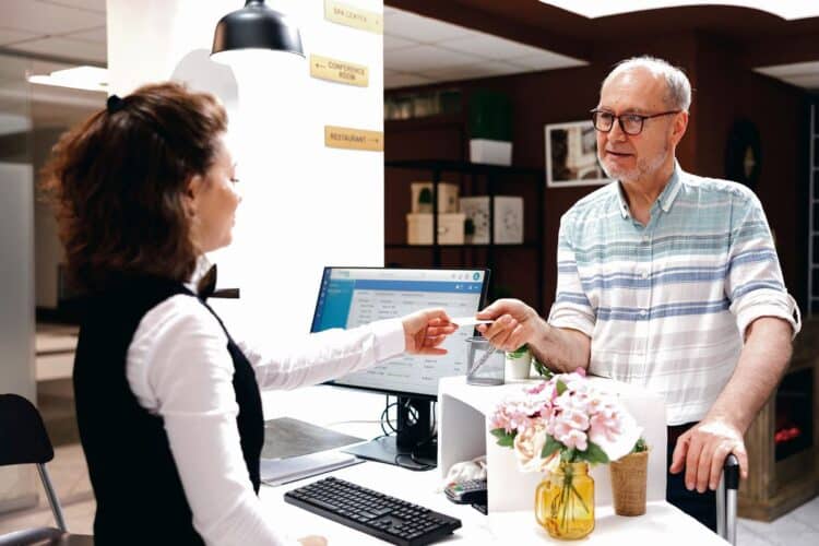 Detailed image shows caucasian retired senior man receiving access card from female receptionist at registration counter. Elderly male tourist giving room key to employee at front desk in hotel lobby.